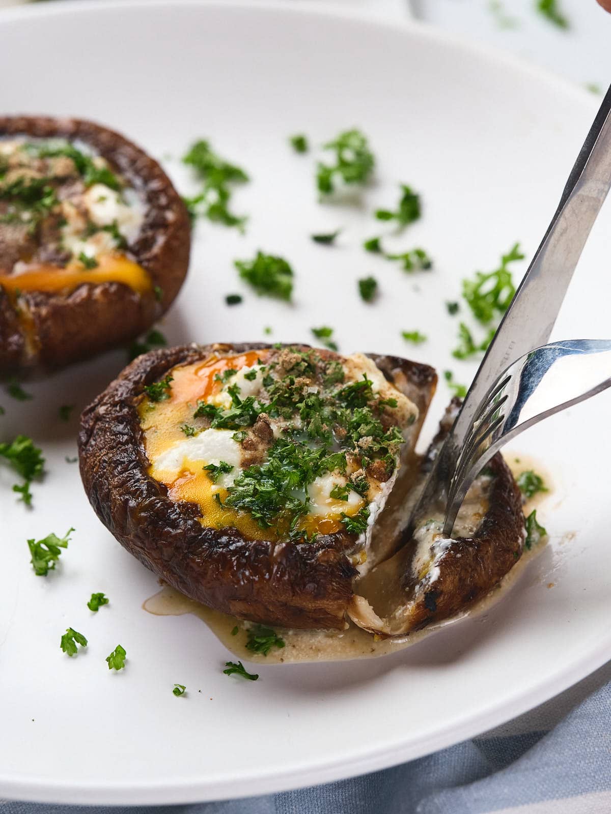 A fork slices into an egg in mushroom garnished with chopped parsley on a white plate.