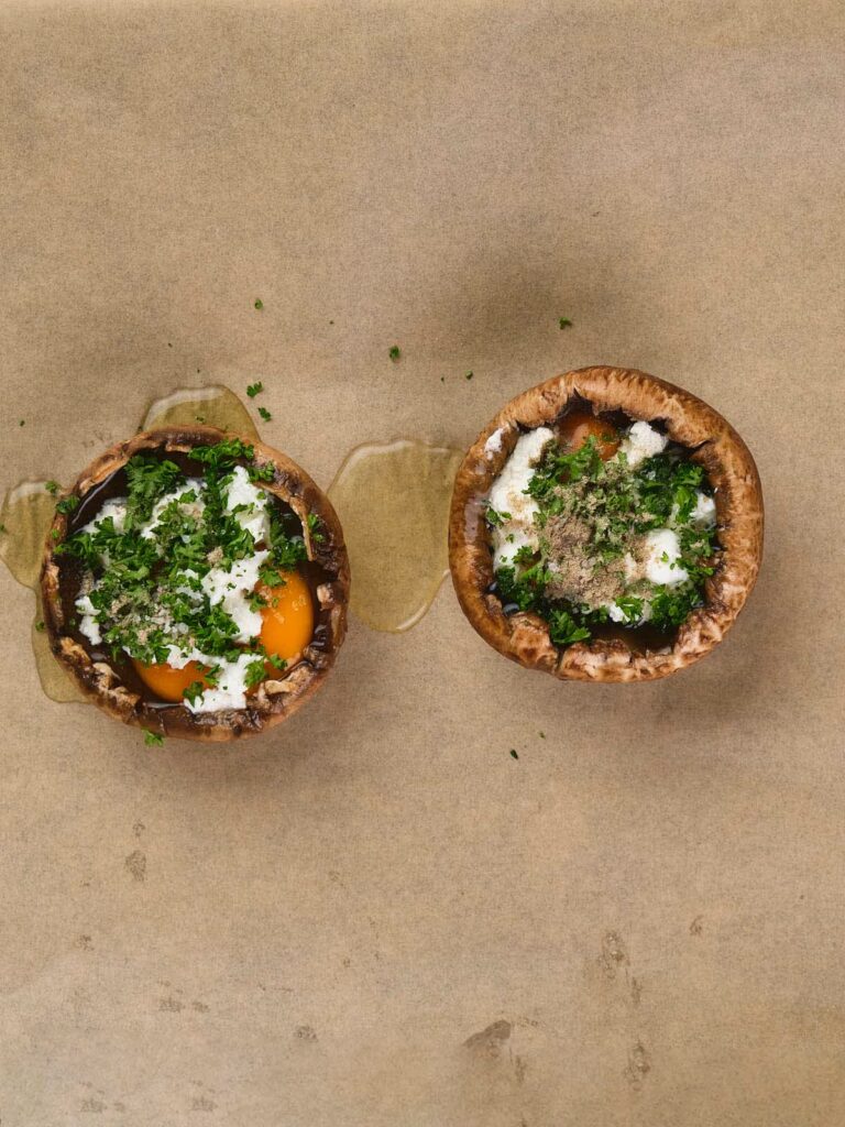 Two mushroom caps filled with eggs, herbs, and cheese are placed on parchment paper.