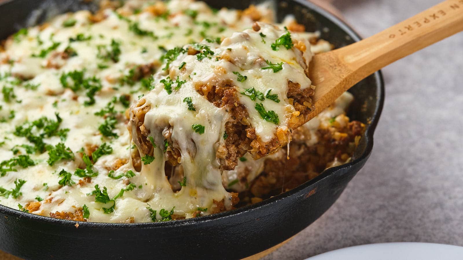 A wooden spoon lifting a serving of italian ground beef skillet garnished with parsley from a cast-iron skillet.