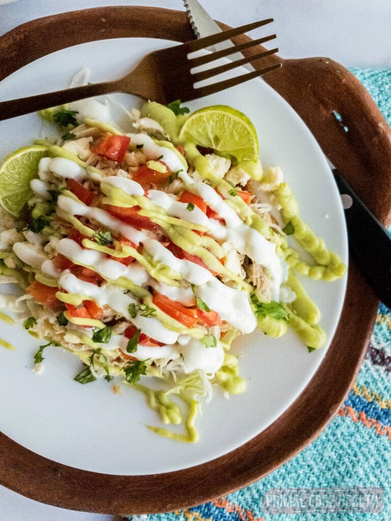 A white plate with a serving of salad topped with chicken, diced tomatoes, lime wedges, and drizzled with white and green sauces. A fork and knife are placed next to the plate on a brown mat.