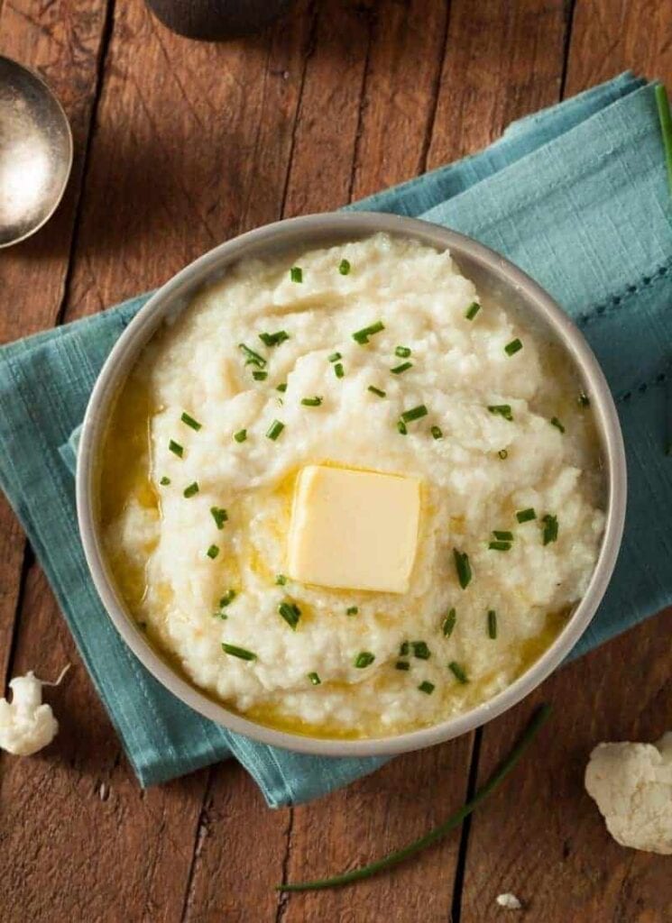 A bowl of mashed cauliflower topped with a pat of butter and garnished with chopped chives, placed on a wooden table with a blue cloth napkin beside it.