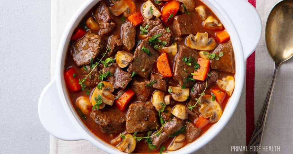 A white bowl filled with beef stew containing chunks of beef, carrots, mushrooms, and topped with herbs. A spoon is placed on a cloth next to the bowl.