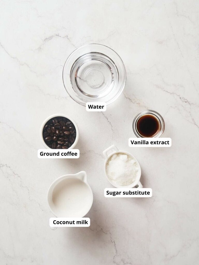 Top-down view of water, vanilla extract, ground coffee, sugar substitute, and coconut milk on a marble surface.