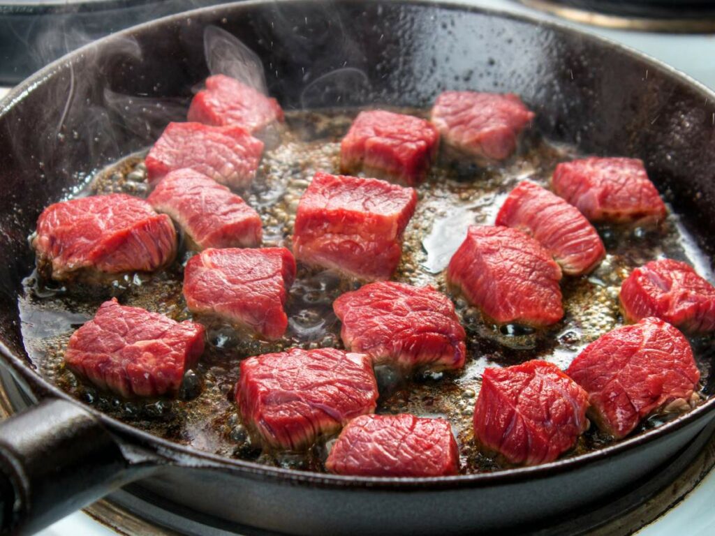 Raw beef cubes sizzling in a hot black skillet, with steam rising from the cooking meat.