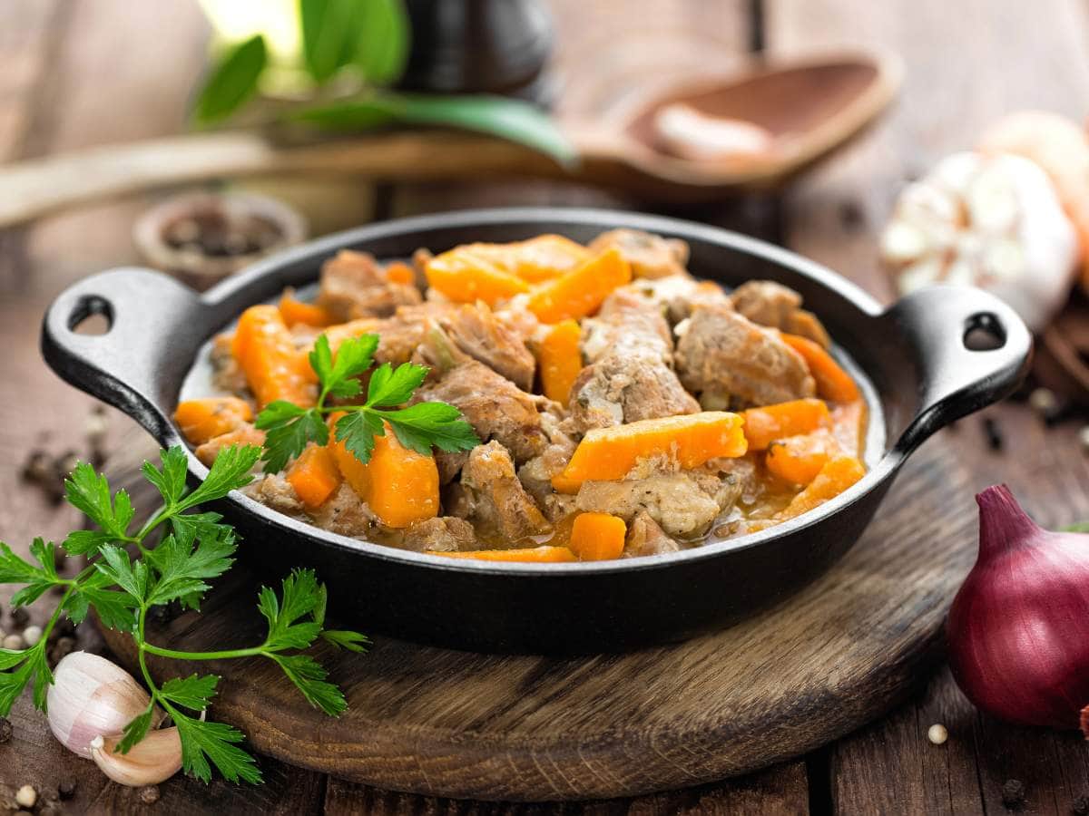 A cast iron pan filled with a stew of meat and carrots, garnished with fresh parsley, sits on a wooden table.