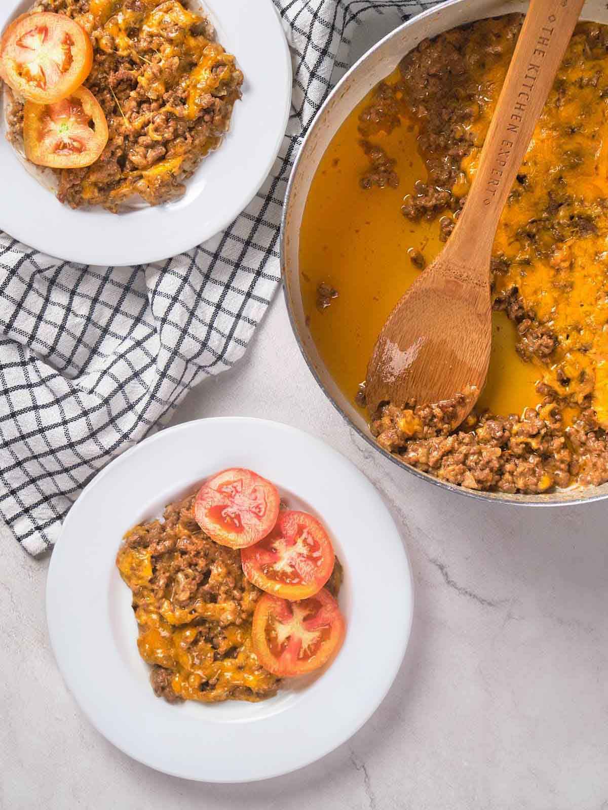 Two plates of seasoned ground beef with sliced tomatoes next to a pan with more of the dish and a wooden spoon.