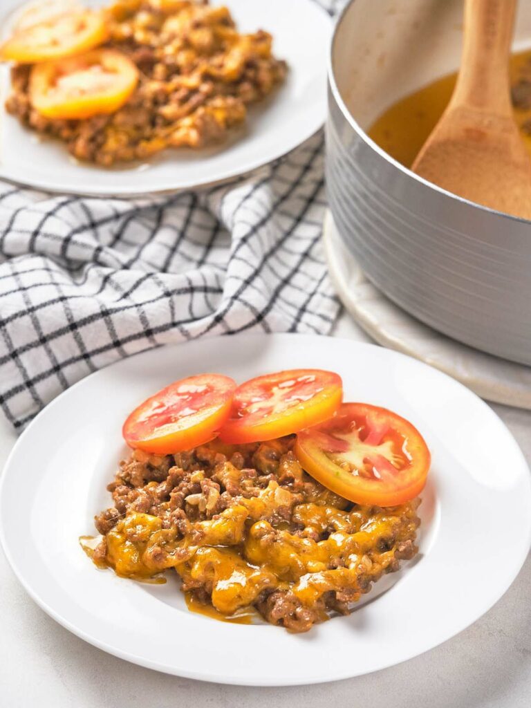 Plate of cheesy ground beef topped with tomato slices, with a checkered cloth and a pot in the background.