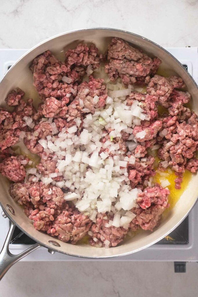 Ground beef and diced onions cooking in a frying pan on a stovetop.