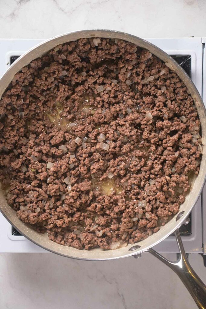 A skillet on a stove contains cooked ground beef with chopped onions.