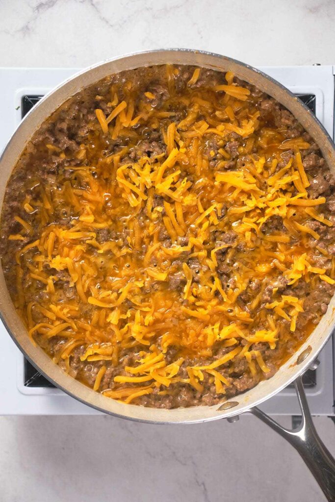 A skillet on a stove contains cooked ground beef with melted shredded cheddar cheese on top.