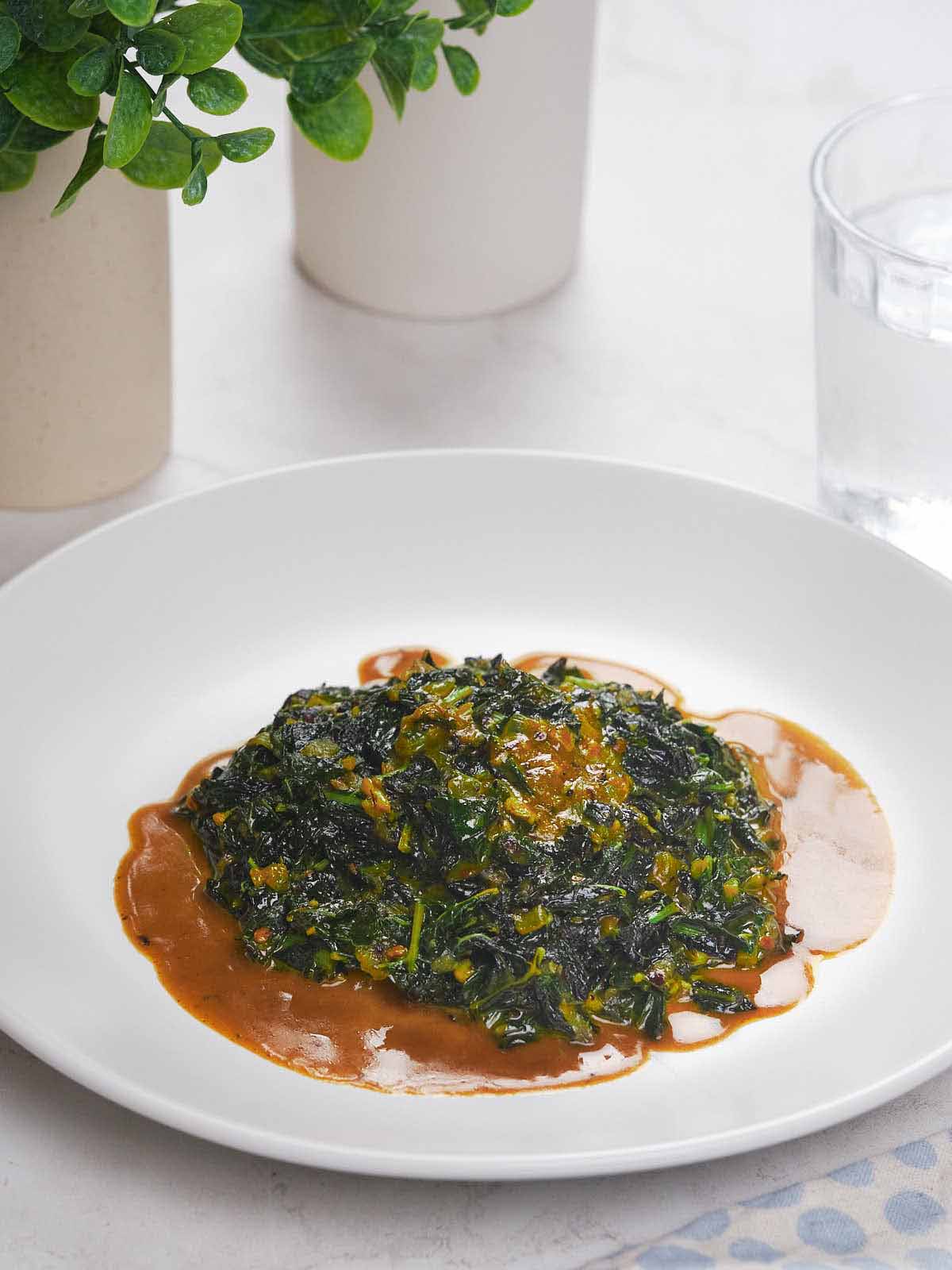 A plate of collard greens in a rich sauce, served on a white dish, with a glass of water and potted plants in the background.