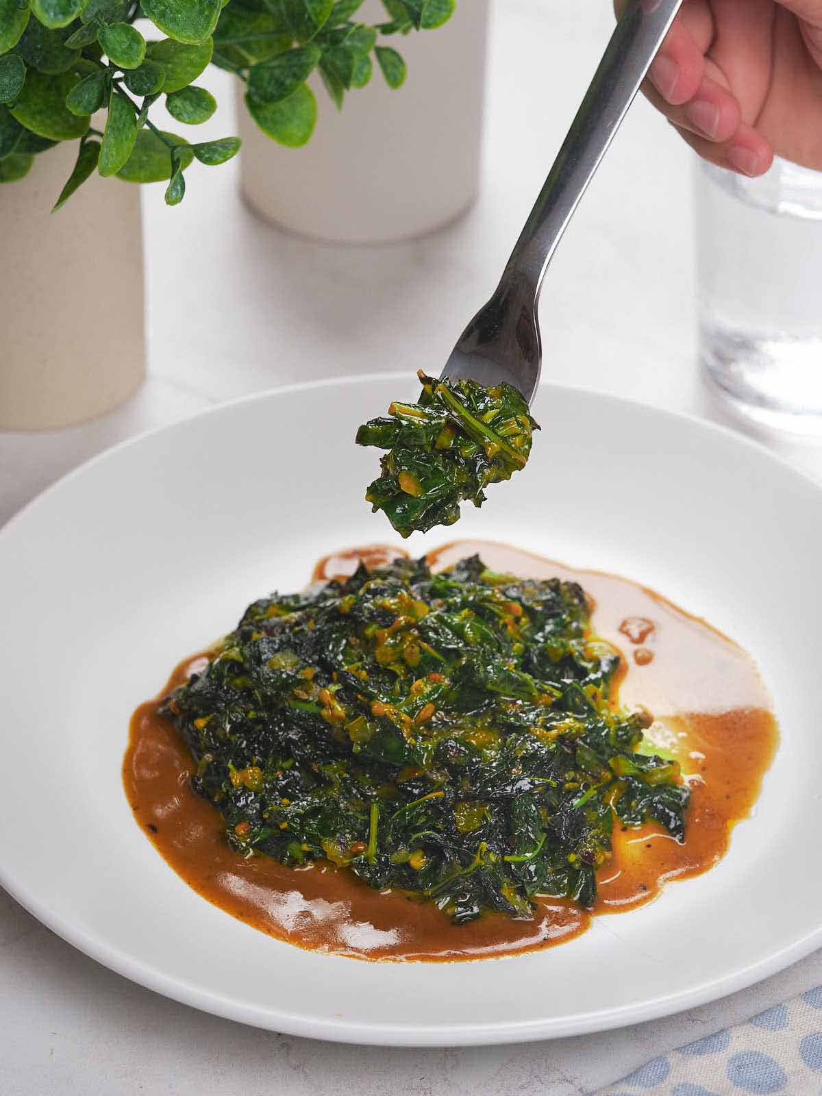 A fork holds cooked collard greens above a plate with greens in a brown sauce.