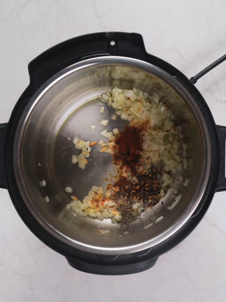 An overhead view of chopped onions and spices in a pressure cooker on a white surface.