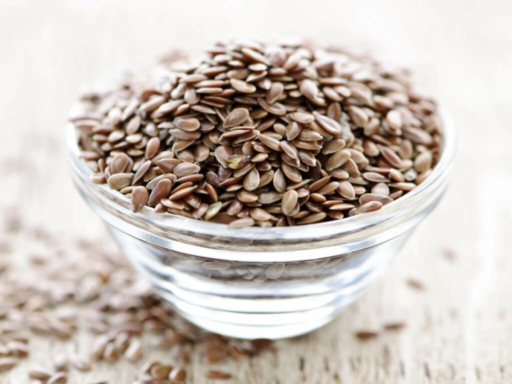 A glass bowl filled with flaxseeds is placed on a wooden surface.