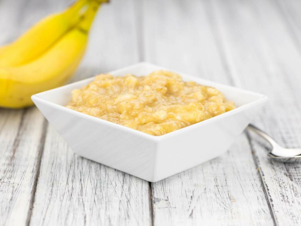 White square bowl of mashed bananas on a wooden surface.