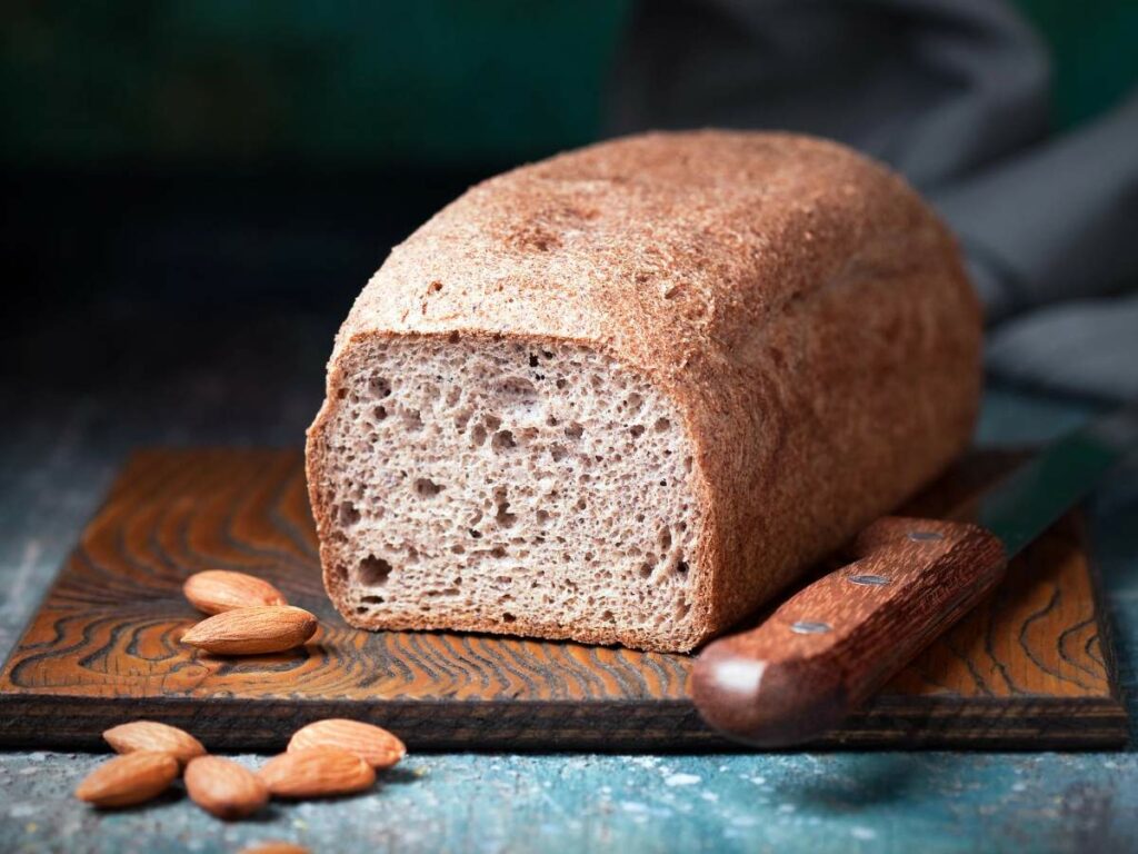 A loaf of brown bread on a wooden cutting board with a knife and almonds beside it.