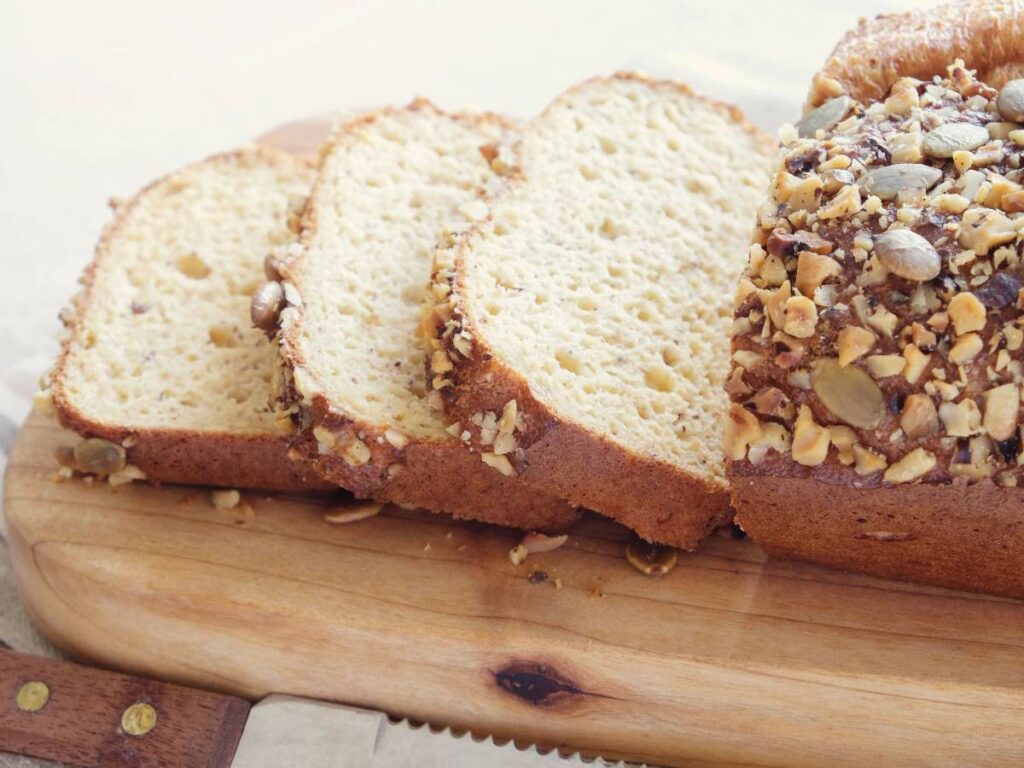 Sliced loaf of bread with nuts and seeds on top, placed on a wooden cutting board.