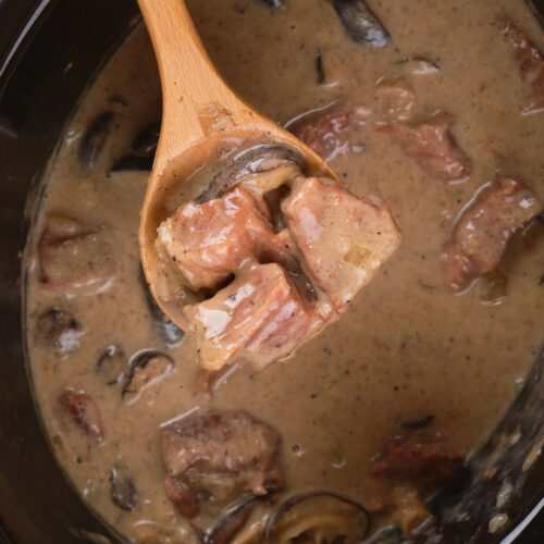 Wooden spoon lifting slow cooker stroganoff with mushrooms from a pot of creamy sauce.