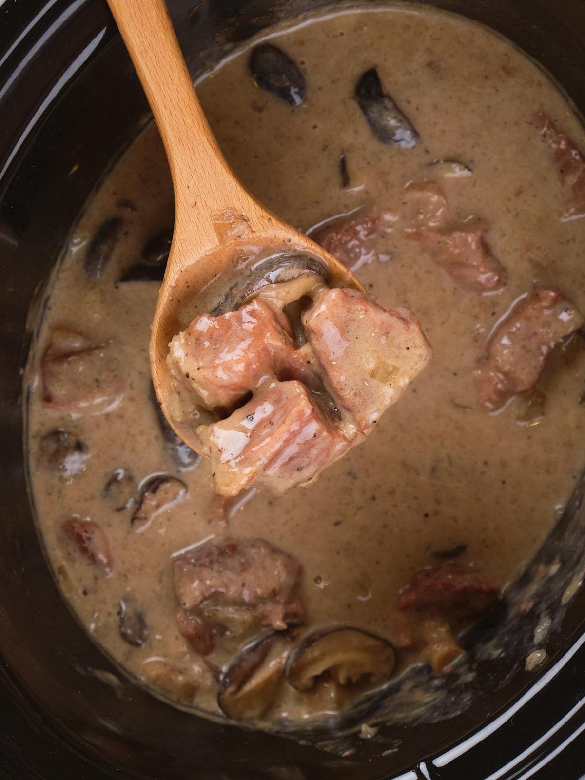 Wooden spoon lifting slow cooker stroganoff with mushrooms from a pot of creamy sauce.