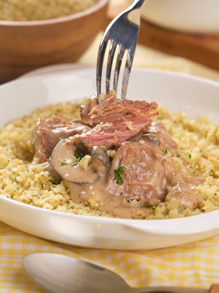 A fork pulls apart tender meat served on a bed of grains, topped with a creamy mushroom sauce and garnished with herbs.