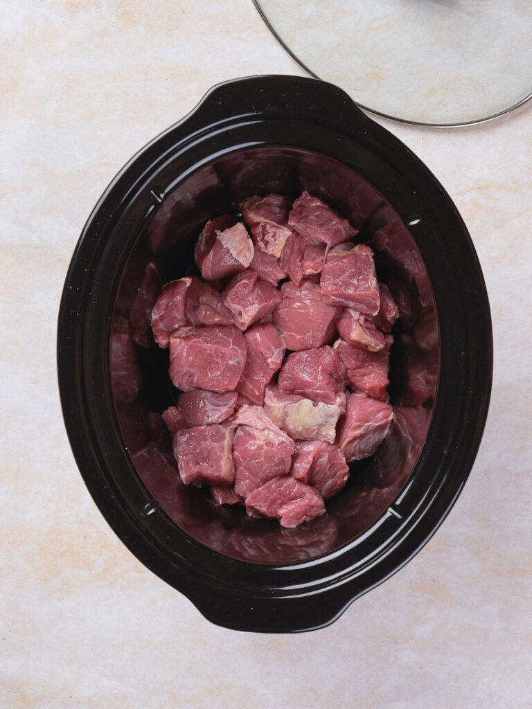 Cubed raw meat placed in a black slow cooker on a light beige countertop.