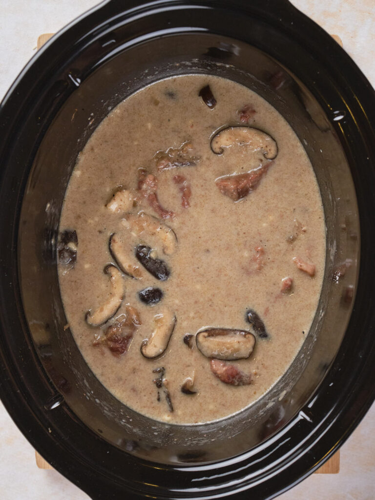 Slow cooker with creamy mushroom and bacon soup containing visible slices of mushrooms and bacon pieces.