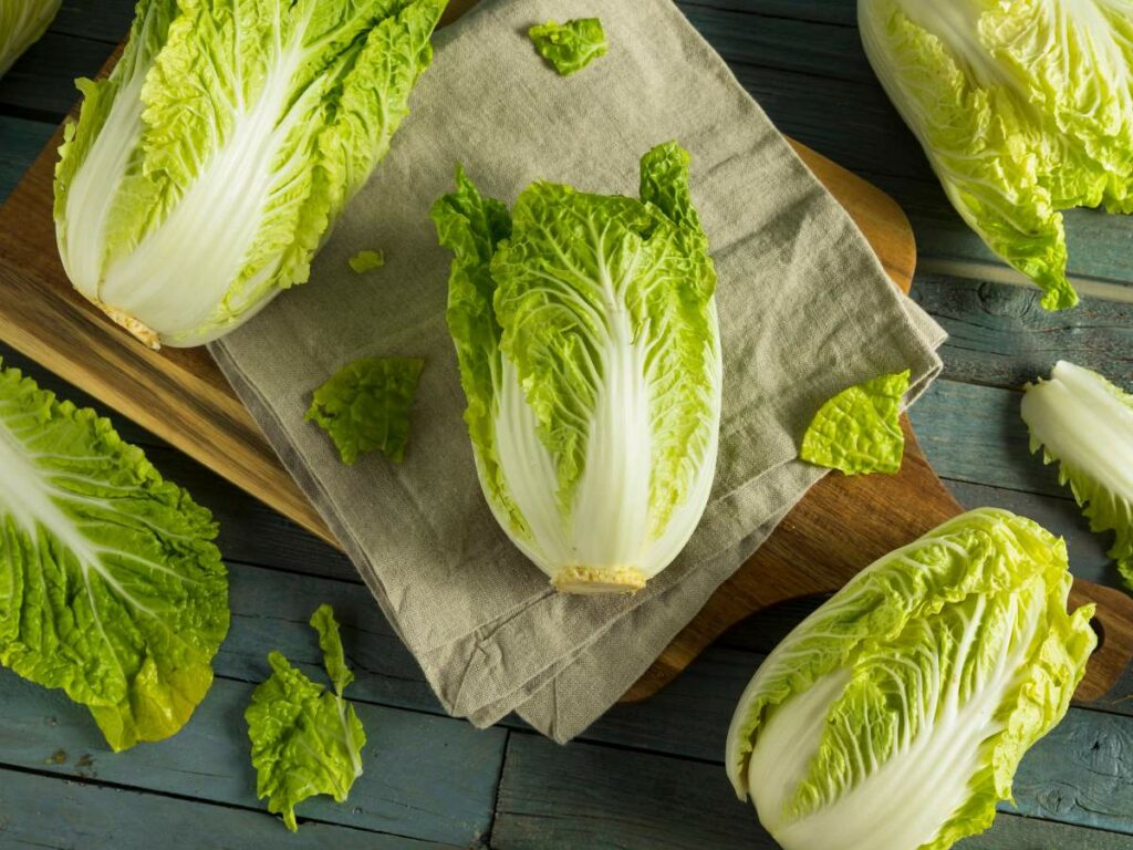 Cabbages on a wooden table, some on a cutting board and cloth, with a few loose green leaves scattered around.