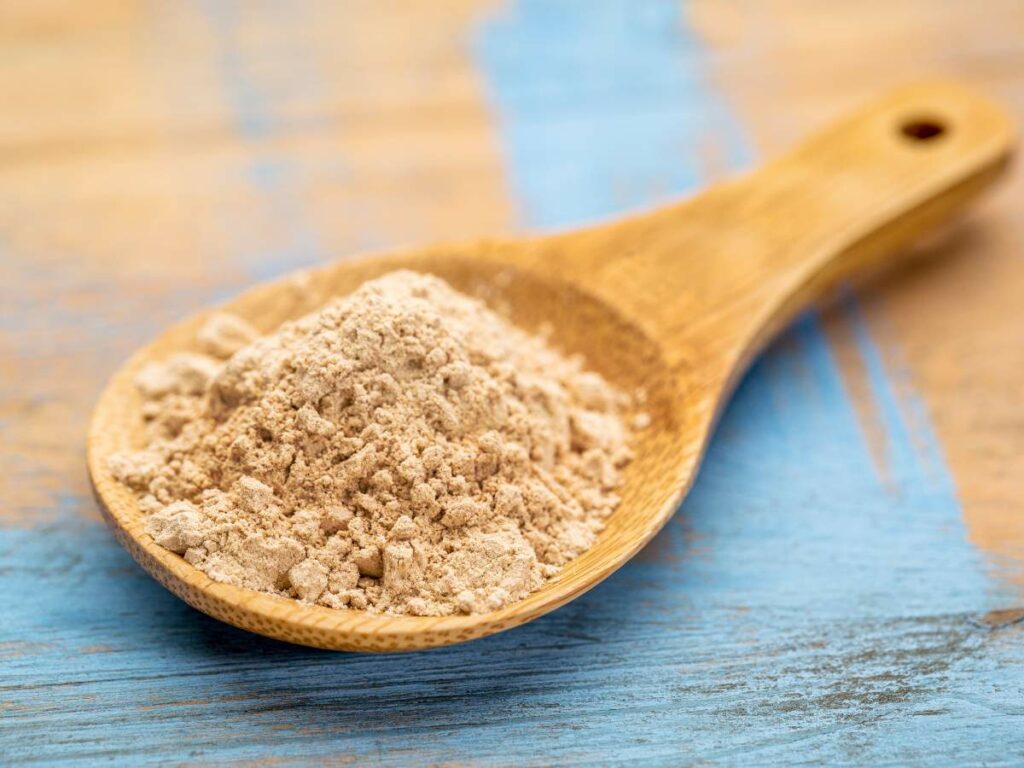 A wooden spoon holds a mound of beige powder on a blue and wooden surface.