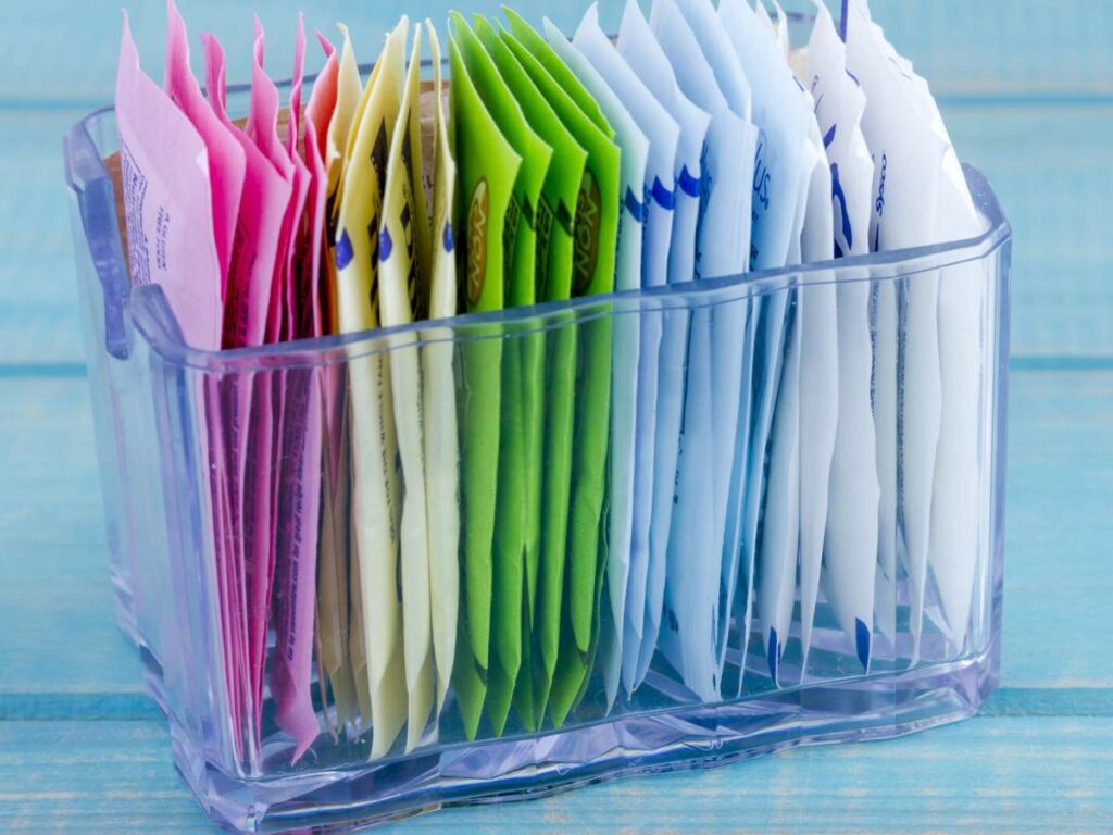 A clear container holds assorted packets of sweeteners in pink, yellow, green, blue, and white.