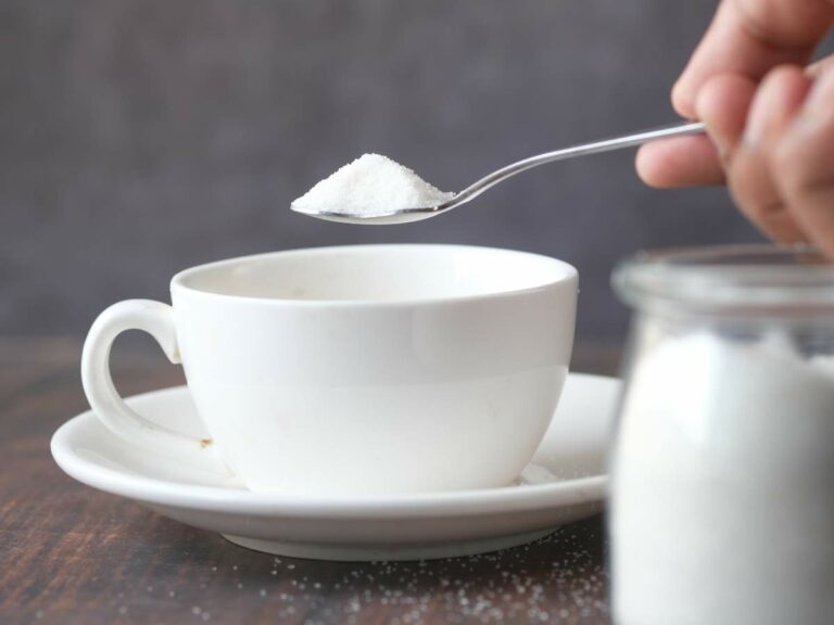 A spoonful of sugar is being added to a white cup on a saucer, with a jar of sugar nearby.