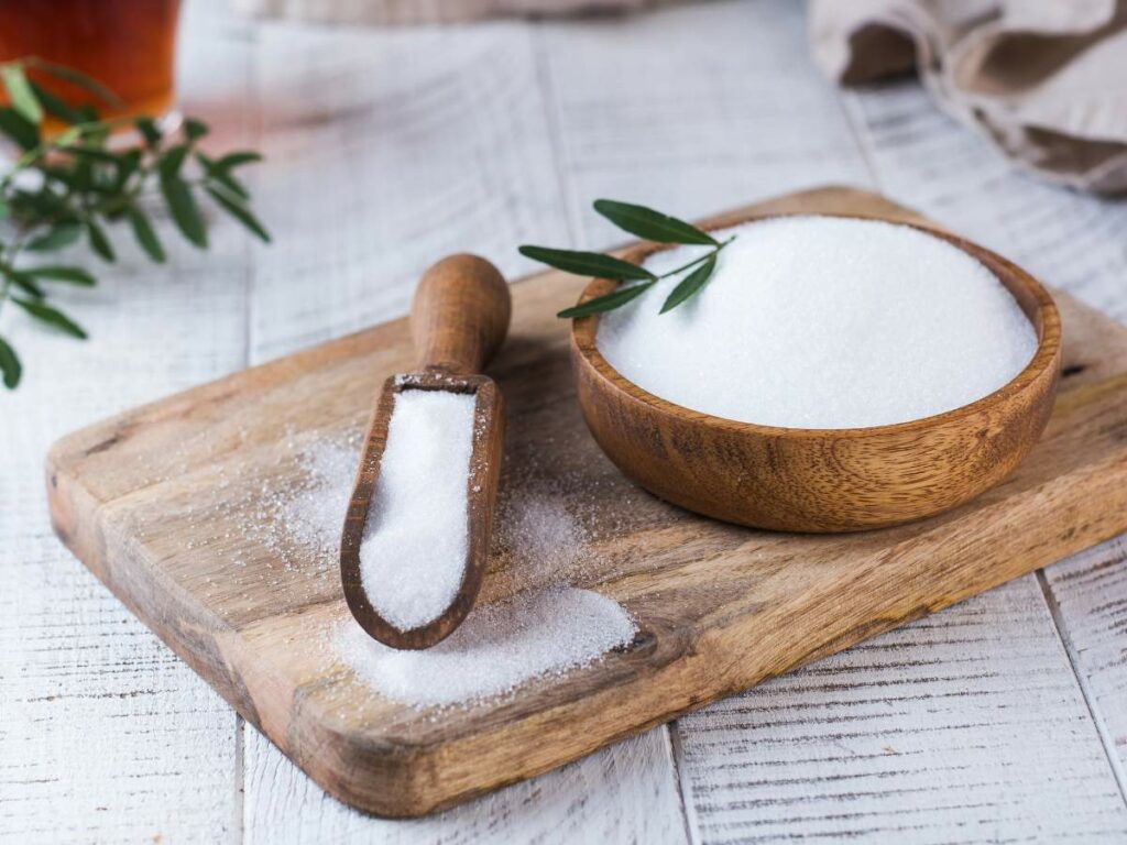 A wooden bowl and scoop filled with white sugar on a wooden cutting board, with a green sprig nearby.