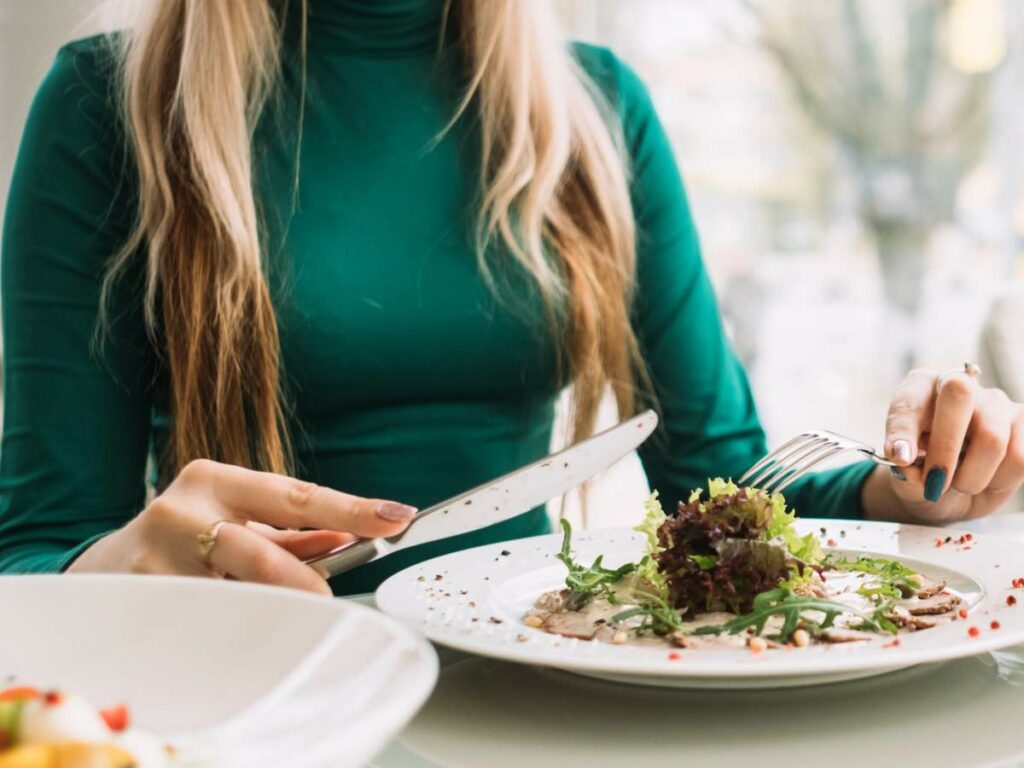 Person in a green top eating a salad with a fork and knife at a dining table.