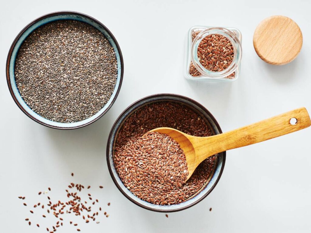 Two bowls and a jar filled with chia and flax seeds, with a wooden spoon resting in a bowl of flax seeds.