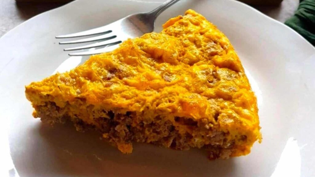 A slice of ground beef and egg casserole with a golden crust on a white plate, accompanied by a fork.