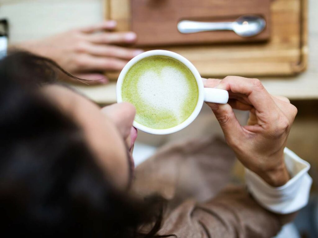 A person holds a cup of green tea latte with heart-shaped foam art, viewed from above.