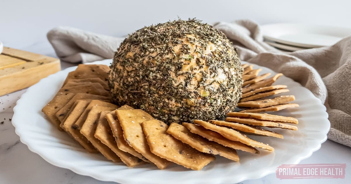 A cheese ball coated with herbs on a white plate, surrounded by crackers.