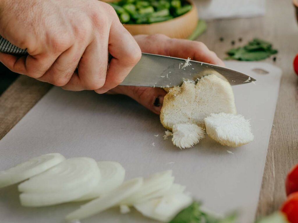 A person slices a mushroom on a cutting board, with sliced onions and herbs nearby.