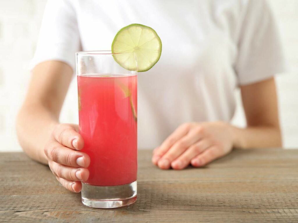 A person in a white shirt holds a glass of pink lemonade with a lime slice on the rim, standing on a wooden table.