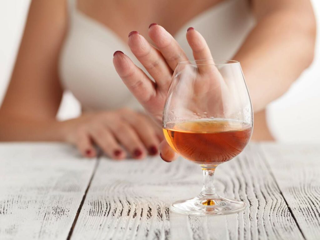 A person in a tank top pushes away a glass of brown liquid on a wooden table.