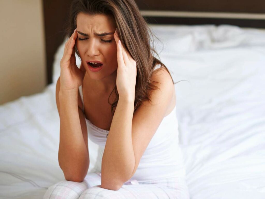 Woman sits on a bed, holding her head with both hands, eyes closed, appearing to have a headache.