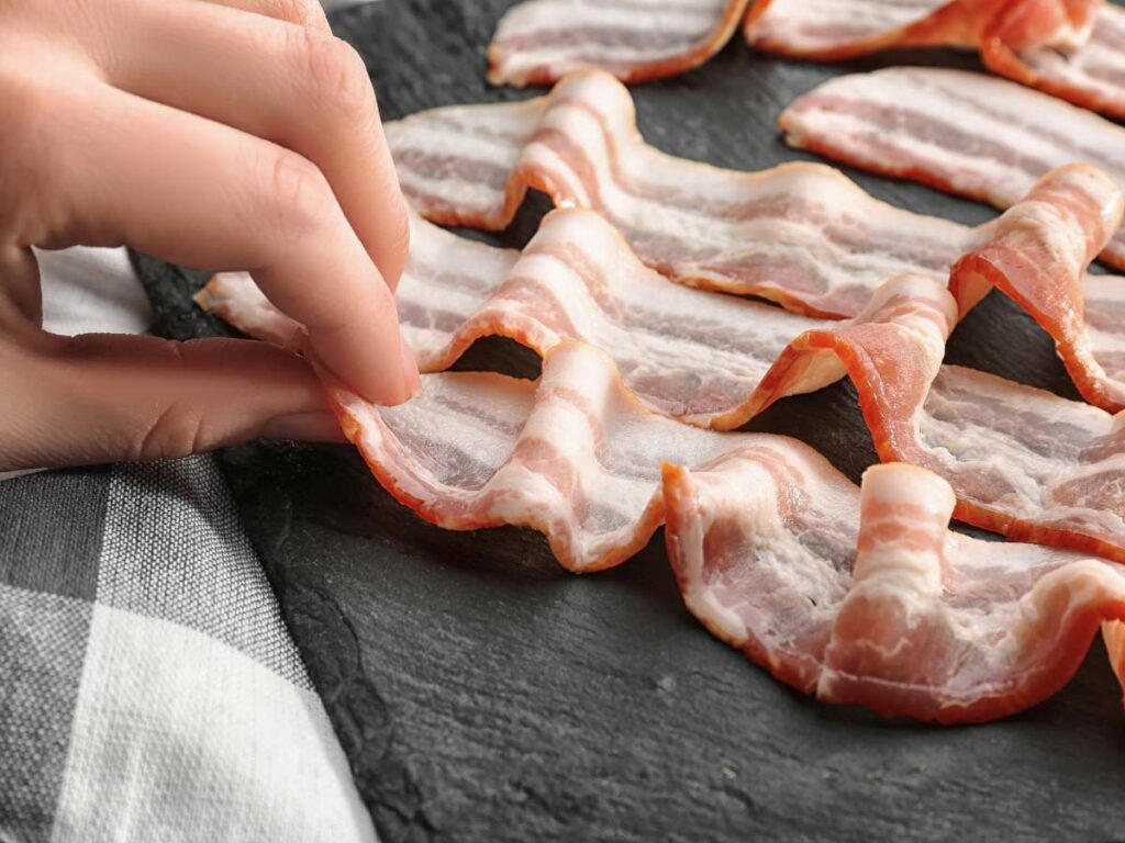 Hand arranging several strips of raw bacon on a dark slate surface.