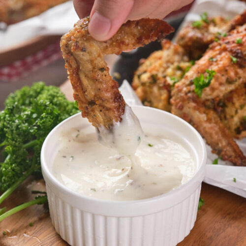 A hand dips a parmesan chicken wing into a small white bowl of creamy sauce.