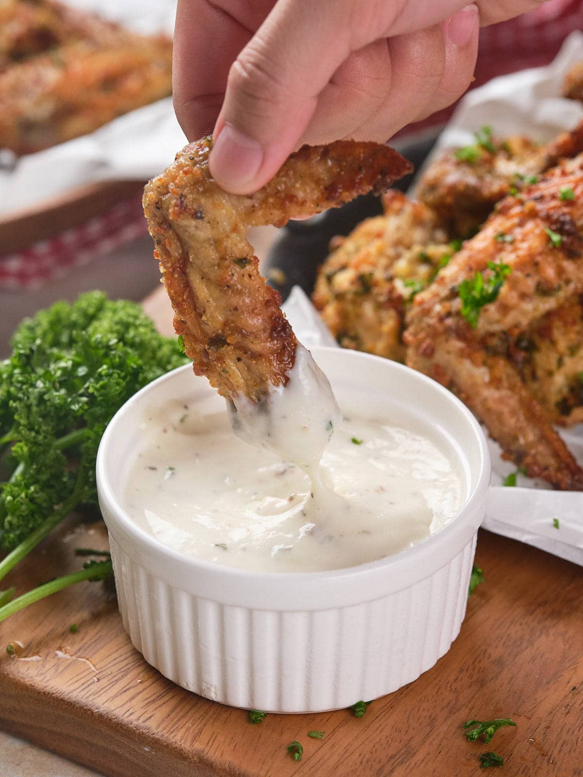 A hand dips a parmesan chicken wing into a small white bowl of creamy sauce.