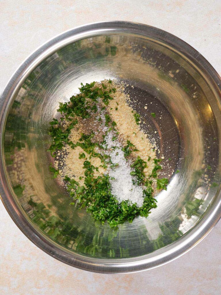 A metal bowl containing a mixture of cheese, chopped parsley, and salt.