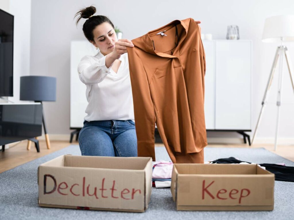 A woman sits on the floor sorting clothes into "Declutter" and "Keep" boxes, holding an orange shirt.