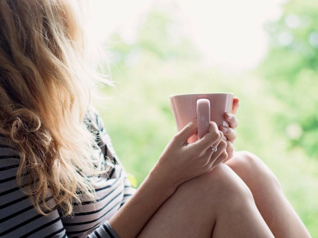 Person with long hair holding a pink mug, sitting by a window.