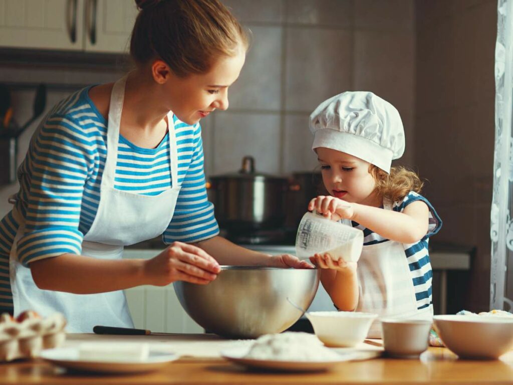 A woman and a child in matching striped shirts and aprons are baking in a kitchen. The child, wearing a chef's hat, pours ingredients into a mixing bowl held by the woman.