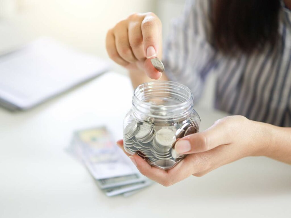 Close-up of a person placing a coin into a glass jar filled with coins.