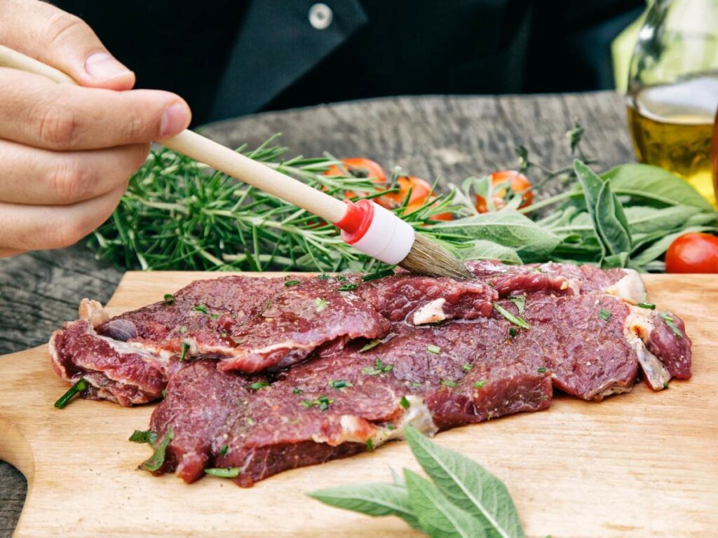 Person brushing raw steaks with oil on a wooden board, surrounded by herbs and tomatoes.