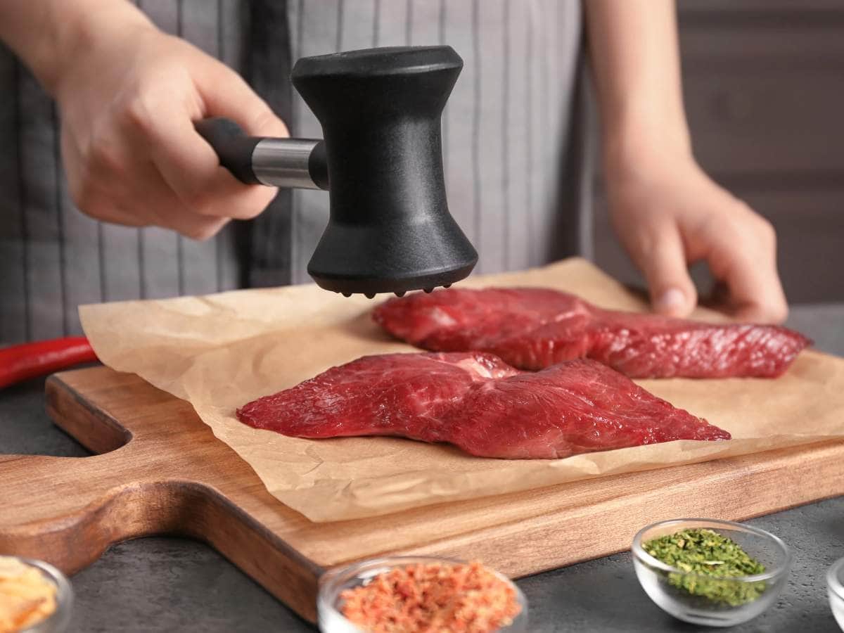 Person tenderizing raw red meat on a wooden board with a meat hammer; spices are in small bowls nearby.